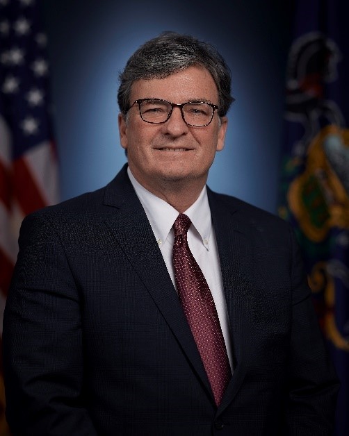 An image of PennDOT Secretary Michael Carroll wearing a black suit jacket, white shirt and maroon tie with a United States flag and a Commonwealth of Pennsylvania flag in the background.